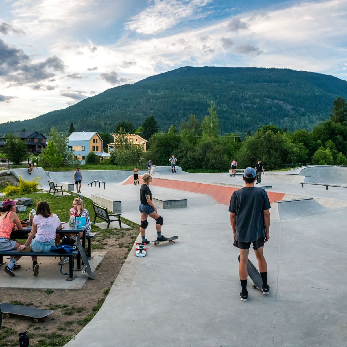 People skateboarding and socializing at a skate park with a mountain backdrop and cloudy sky.
