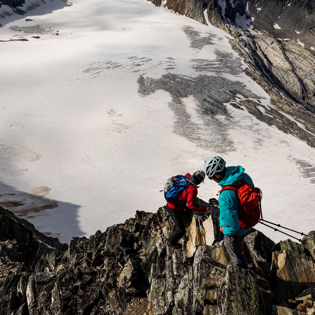 Two climbers wearing helmets and backpacks ascend a rocky ridge with snowy terrain and a small lake visible in the background.