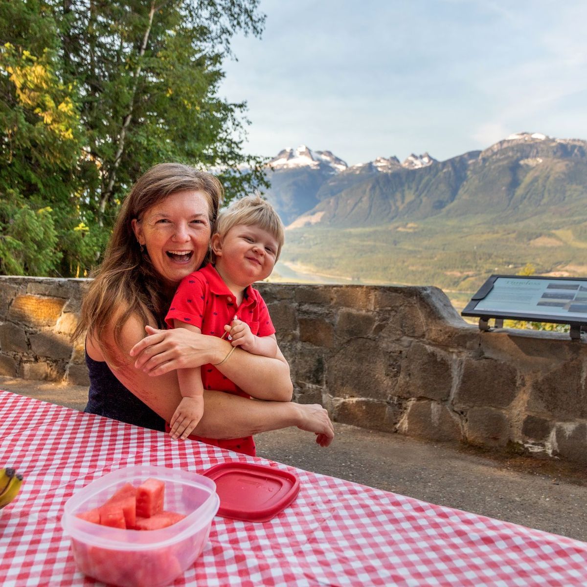 A woman and child smile at a picnic table with bananas and watermelon, set against a scenic mountain backdrop.