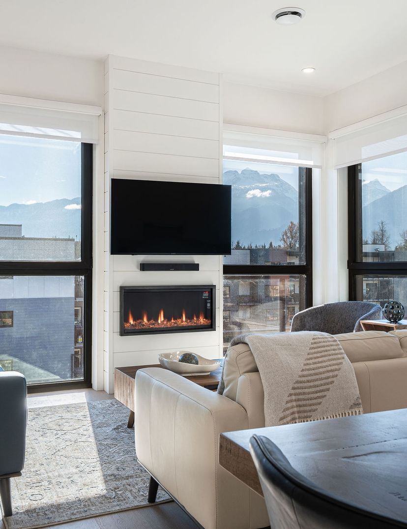 Modern living room with large windows, mountain views, a wall-mounted TV, and a fireplace. Cozy seating and a wooden coffee table complete the space.
