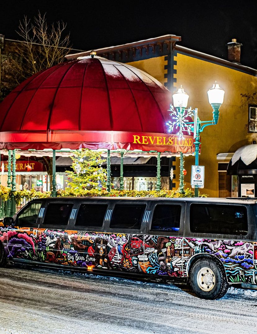 Graffiti-covered stretch limousine parked on a snowy street at night, near buildings with a red dome and lit street lamps.