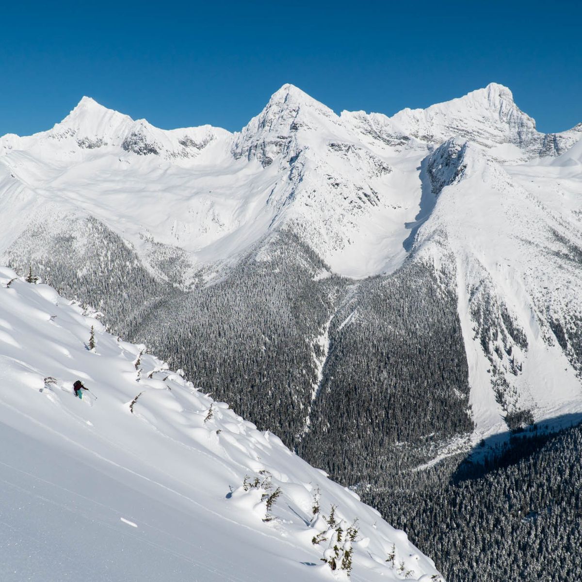 A skier descends a snowy mountain slope with a backdrop of rugged, snow-covered peaks under a clear blue sky.