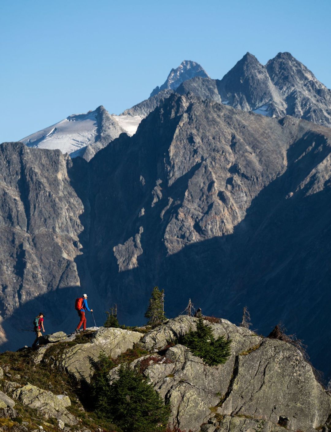 Two hikers in red jackets stand on a rocky ridge with towering, jagged mountains in the background under a clear blue sky.