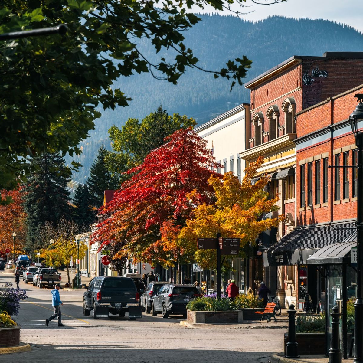 Small town main street in autumn with red and orange trees, brick storefronts, parked cars and a mountain backdrop.