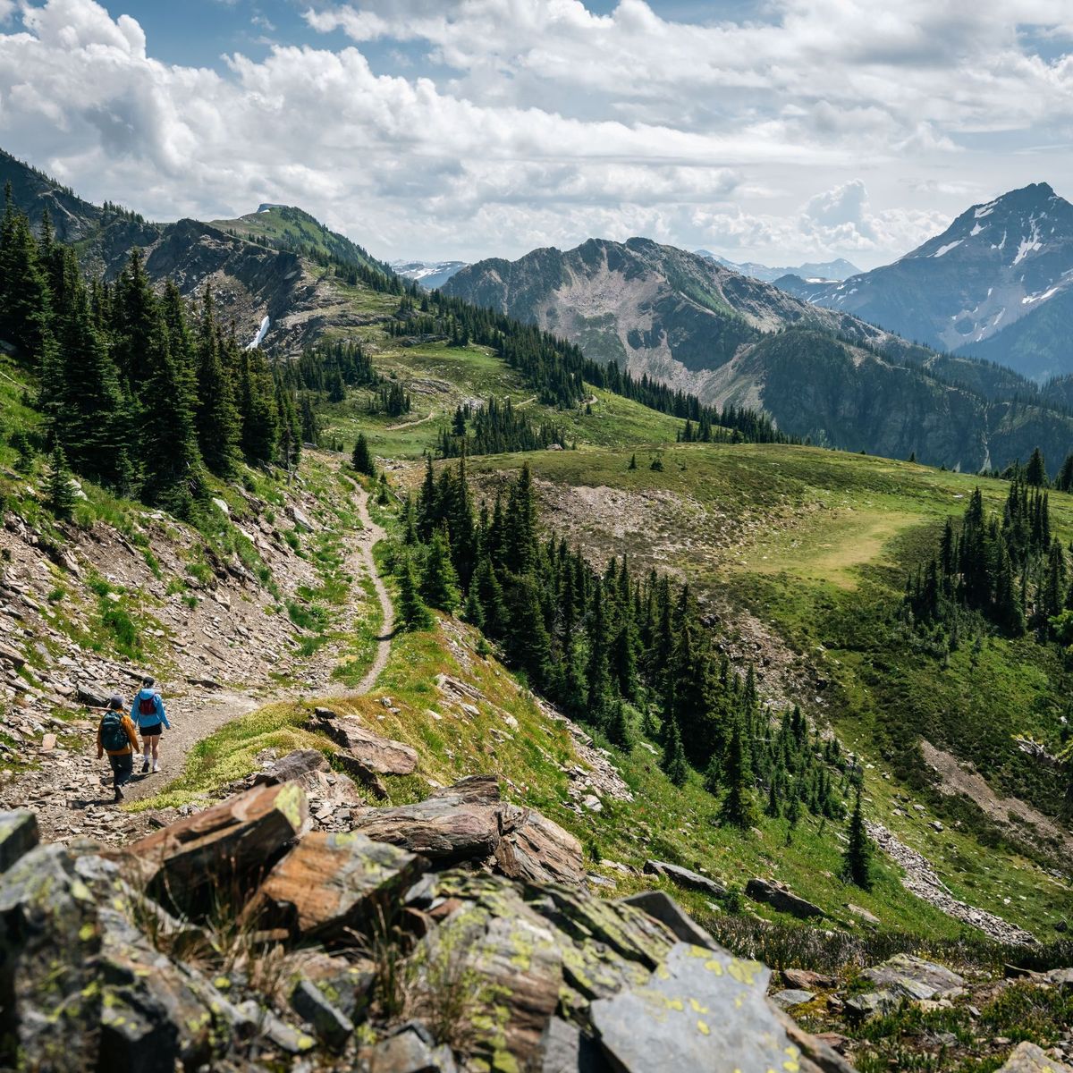Two hikers on a rocky alpine trail winding through green ridges and fir trees toward a distant snow-capped mountain under a cloudy sky.