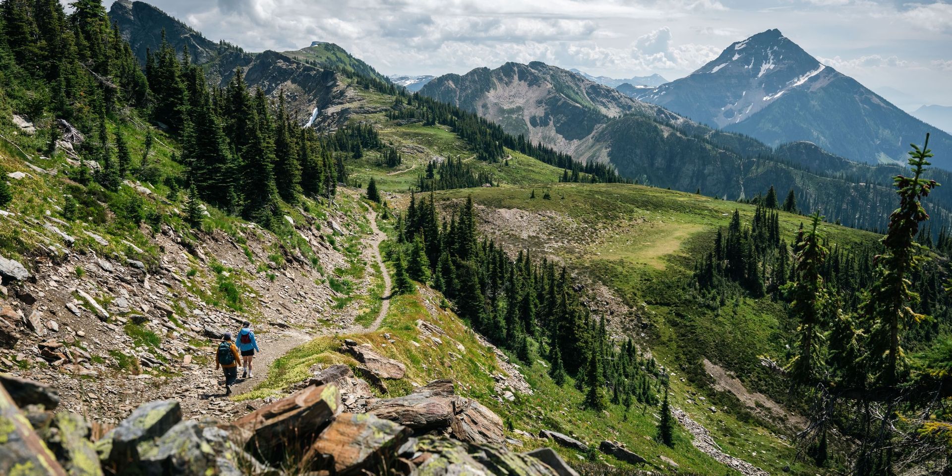 Two hikers on a rocky alpine trail winding through green ridges and fir trees toward a distant snow-capped mountain under a cloudy sky.