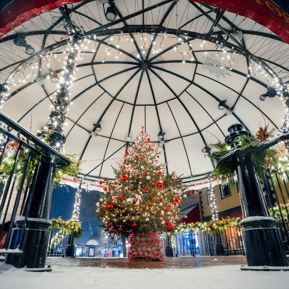Snow-dusted bandstand lit with white string lights, garlands, and a decorated Christmas tree with red ornaments.