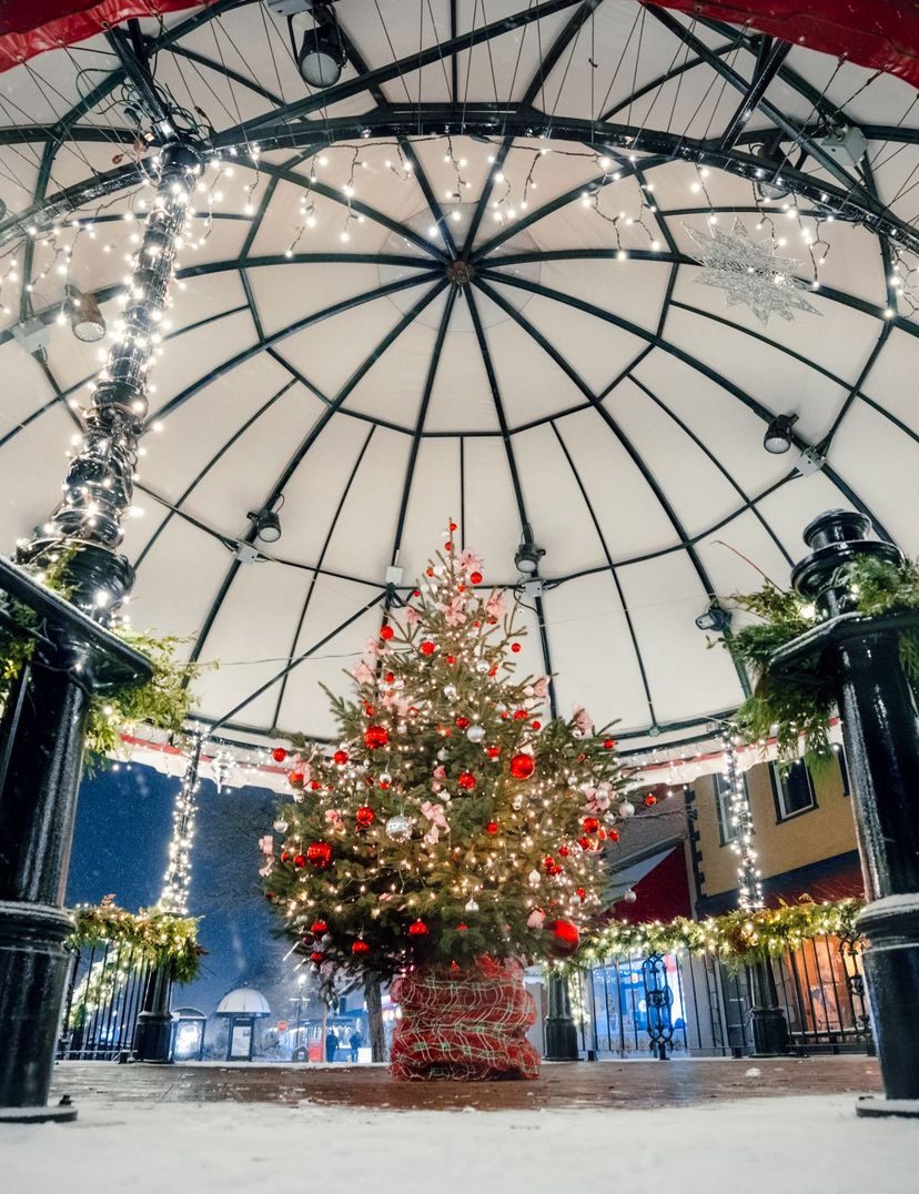 Snow-dusted bandstand lit with white string lights, garlands, and a decorated Christmas tree with red ornaments.