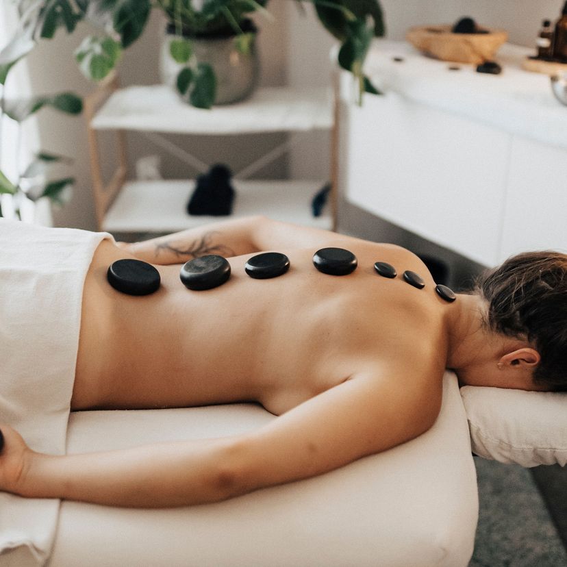 Person lying face down on a massage table with hot stones placed along their back, in a serene spa setting with plants and soft lighting.