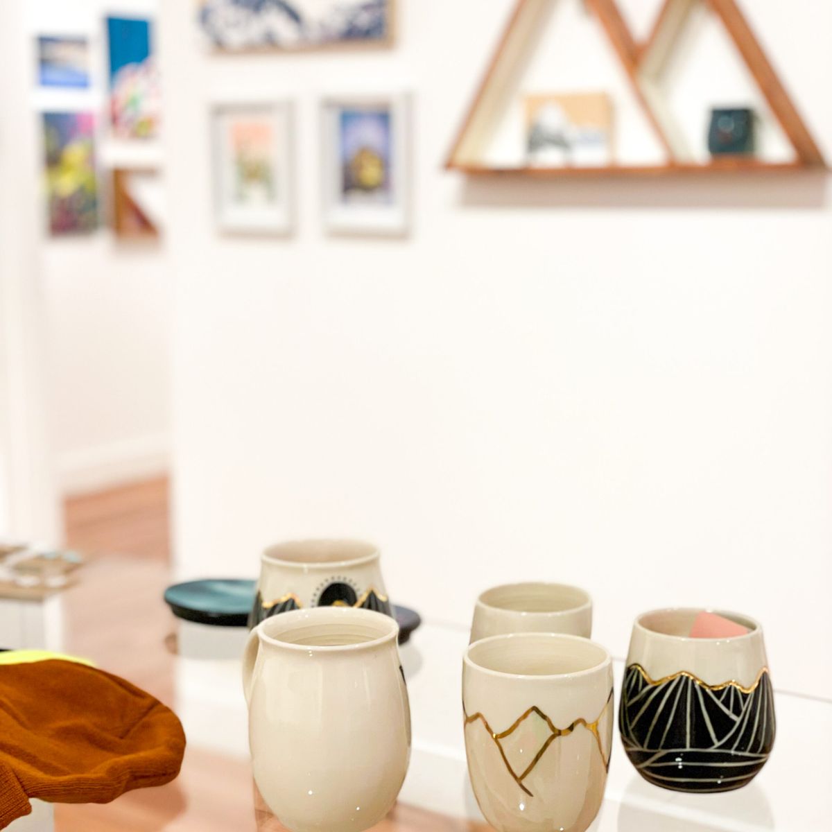 Ceramic mugs with geometric designs on a table in a gallery, with framed artwork and triangular shelves in the background.