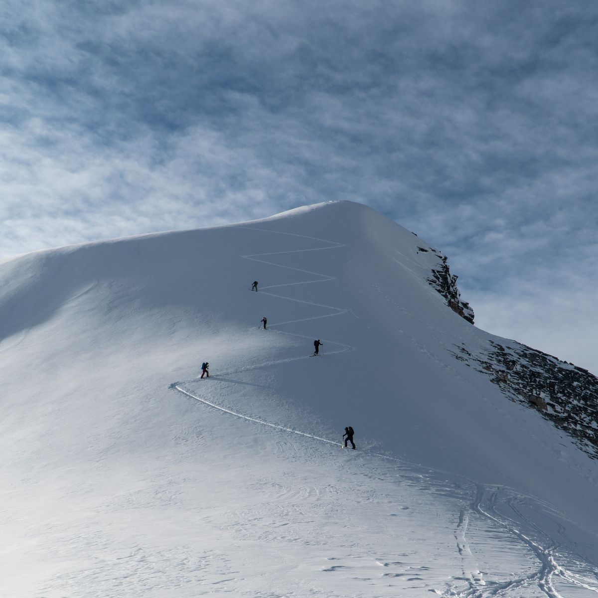 Climbers ascending a snowy mountain slope in a zigzag pattern under a partly cloudy sky.