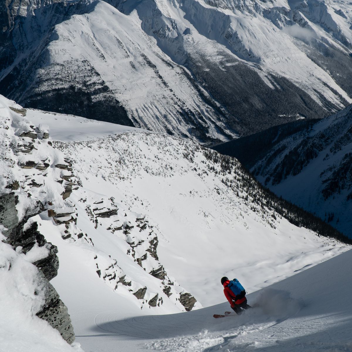 A skier in a red jacket descends a couloir, surrounded by rugged, snow-covered peaks under a clear blue sky.