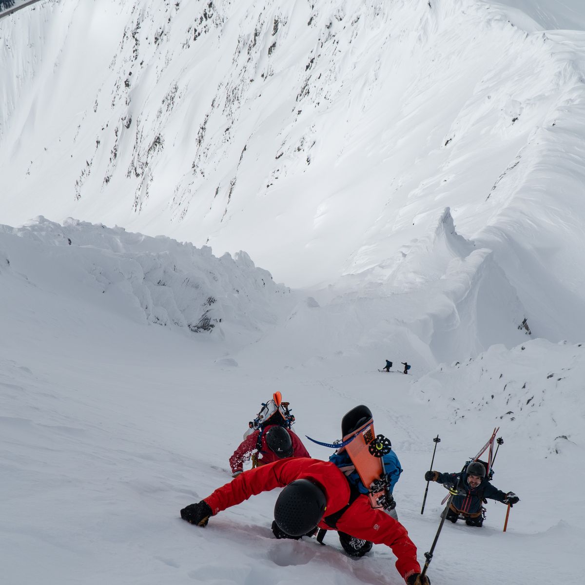Three climbers in winter gear ascend a steep, snowy mountain slope, using ice axes for support, with a mountainous backdrop.
