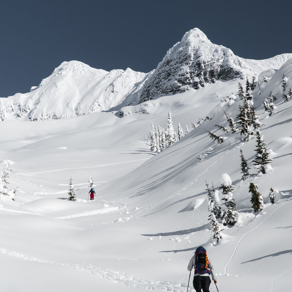 A person skis through a snow-covered mountain landscape under a clear blue sky, with another skier in the distance.