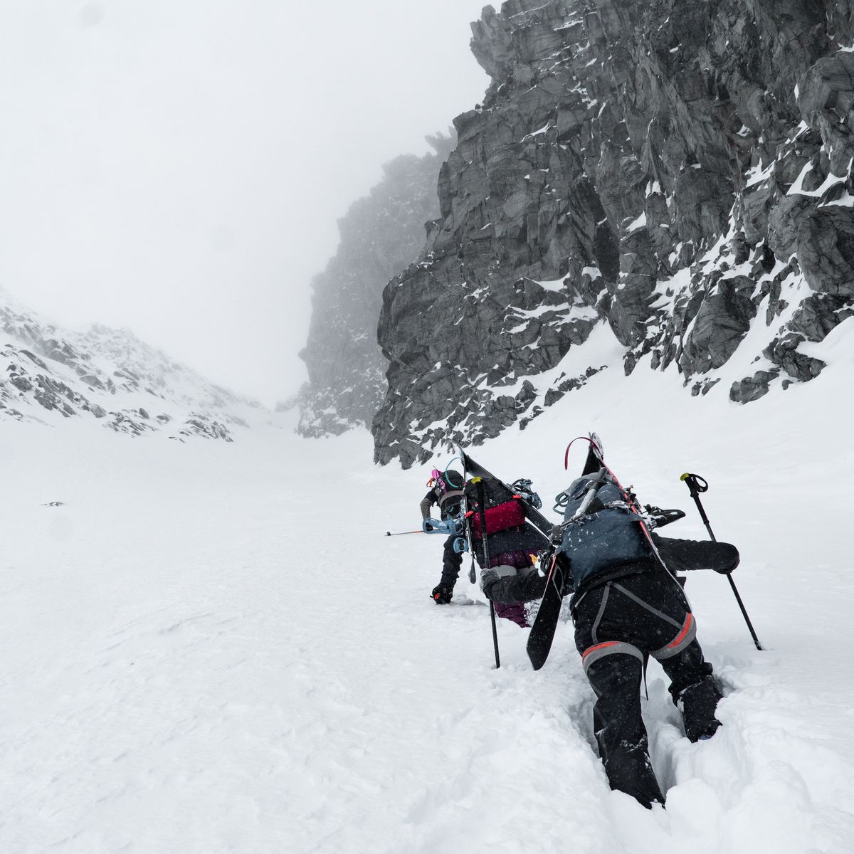 Two climbers with gear ascend a steep, snowy mountain slope, surrounded by rugged rock formations under a cloudy sky.