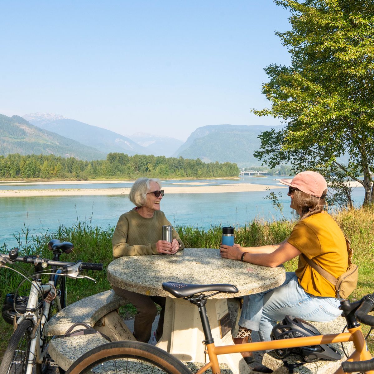 Two people sit at a stone table by a river, surrounded by trees and mountains, with bicycles nearby on a clear day.