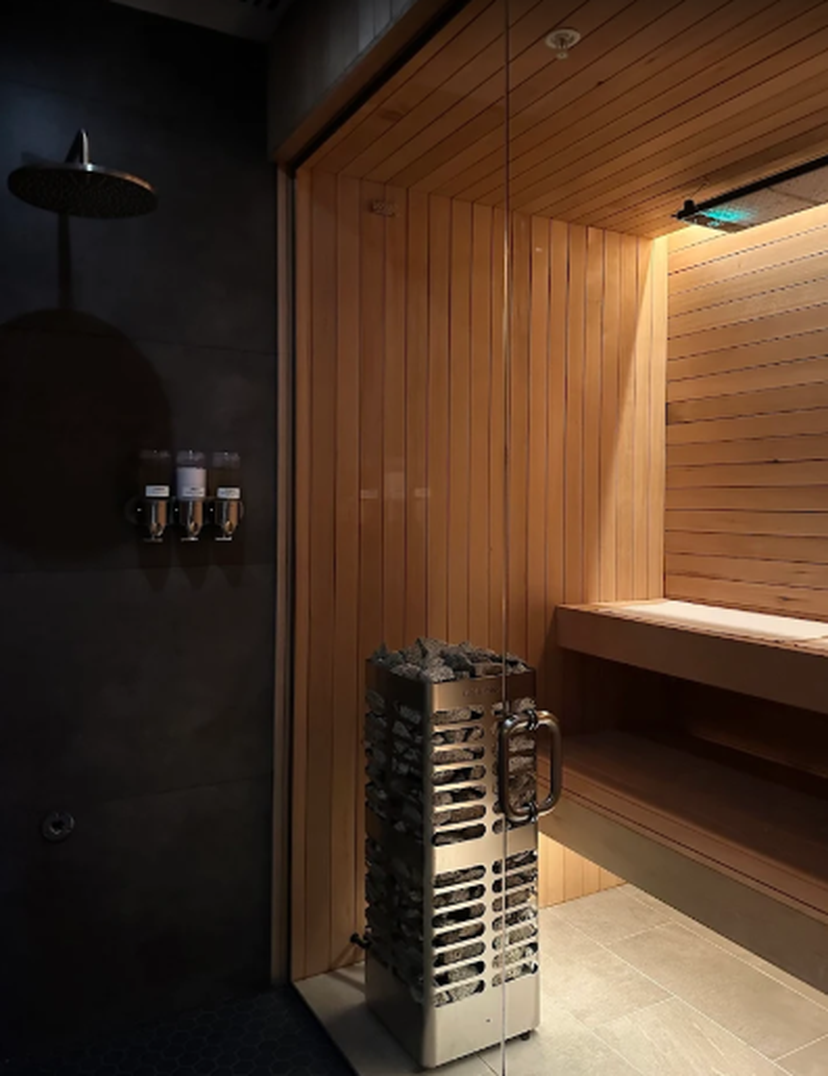 Modern sauna with wooden interior, glass door, bench seating, and a metal heater with stones. Adjacent dark-tiled shower area with overhead showerhead.