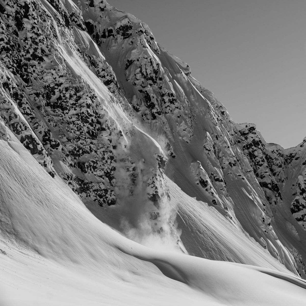 Snow-covered mountain slope with a small avalanche cascading down, surrounded by rugged, rocky peaks under a clear sky. Black and white.