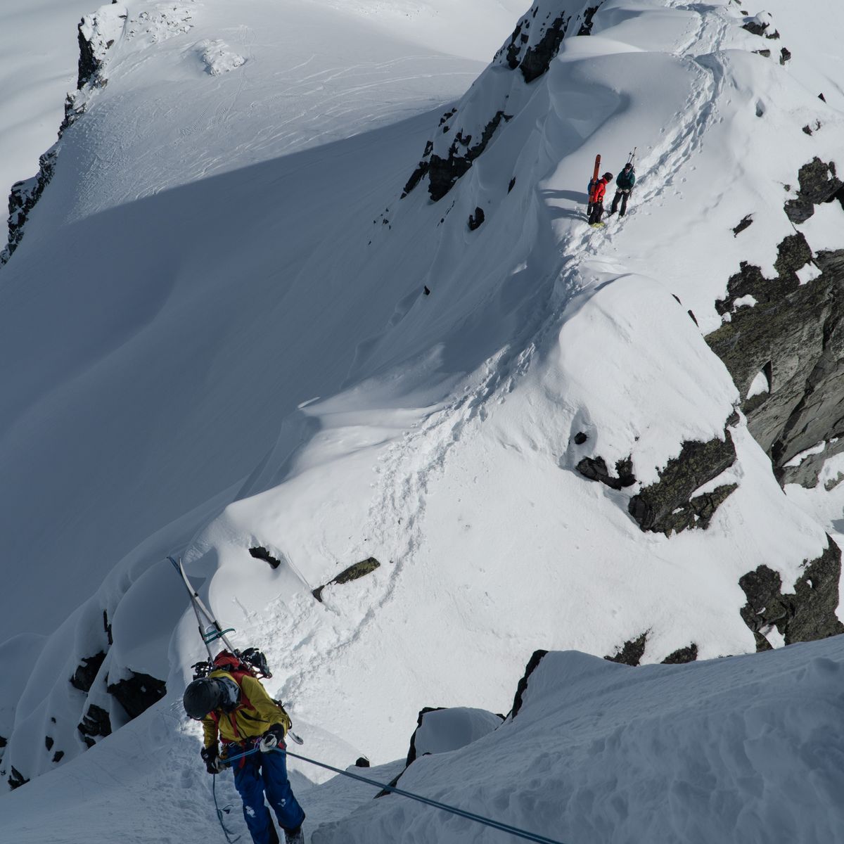 Skiers navigate a narrow, snowy ridge with steep drop-offs, using ropes for safety in a mountainous, winter landscape.
