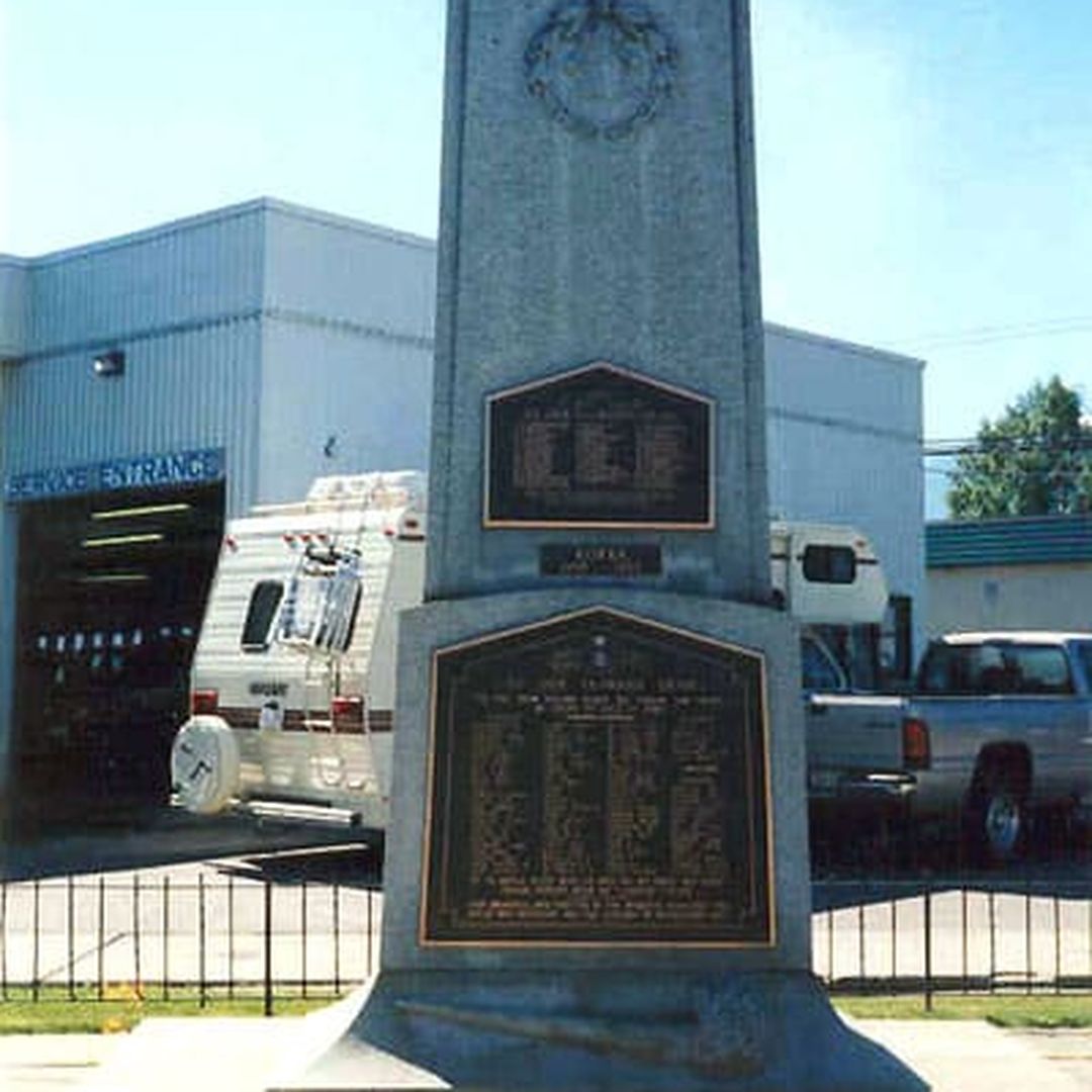 A tall stone memorial with bronze plaques stands in front of a garage, surrounded by orange flowers and parked vehicles.