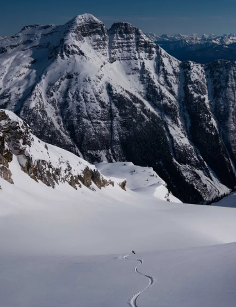 Snowy mountain landscape with a lone skier leaving tracks on a vast, untouched slope. Majestic peaks rise in the background under a clear sky.