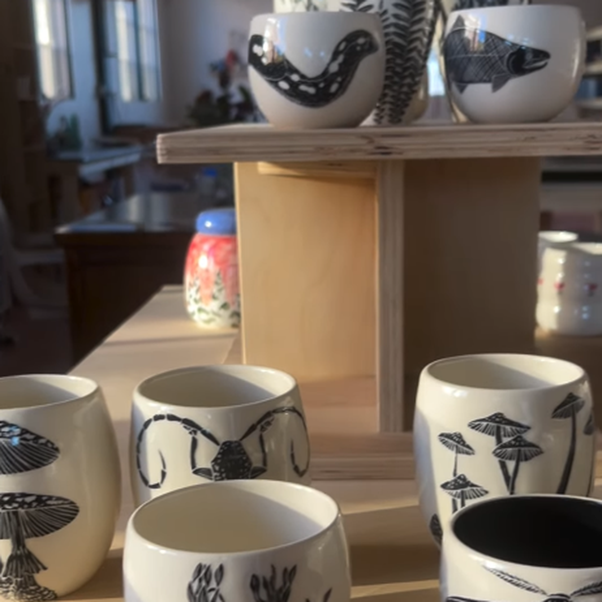 Ceramic cups with black botanical and animal designs displayed on wooden shelves in a sunlit room.