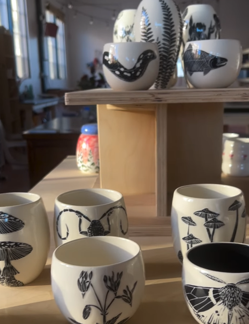 Ceramic cups with black botanical and animal designs displayed on wooden shelves in a sunlit room.