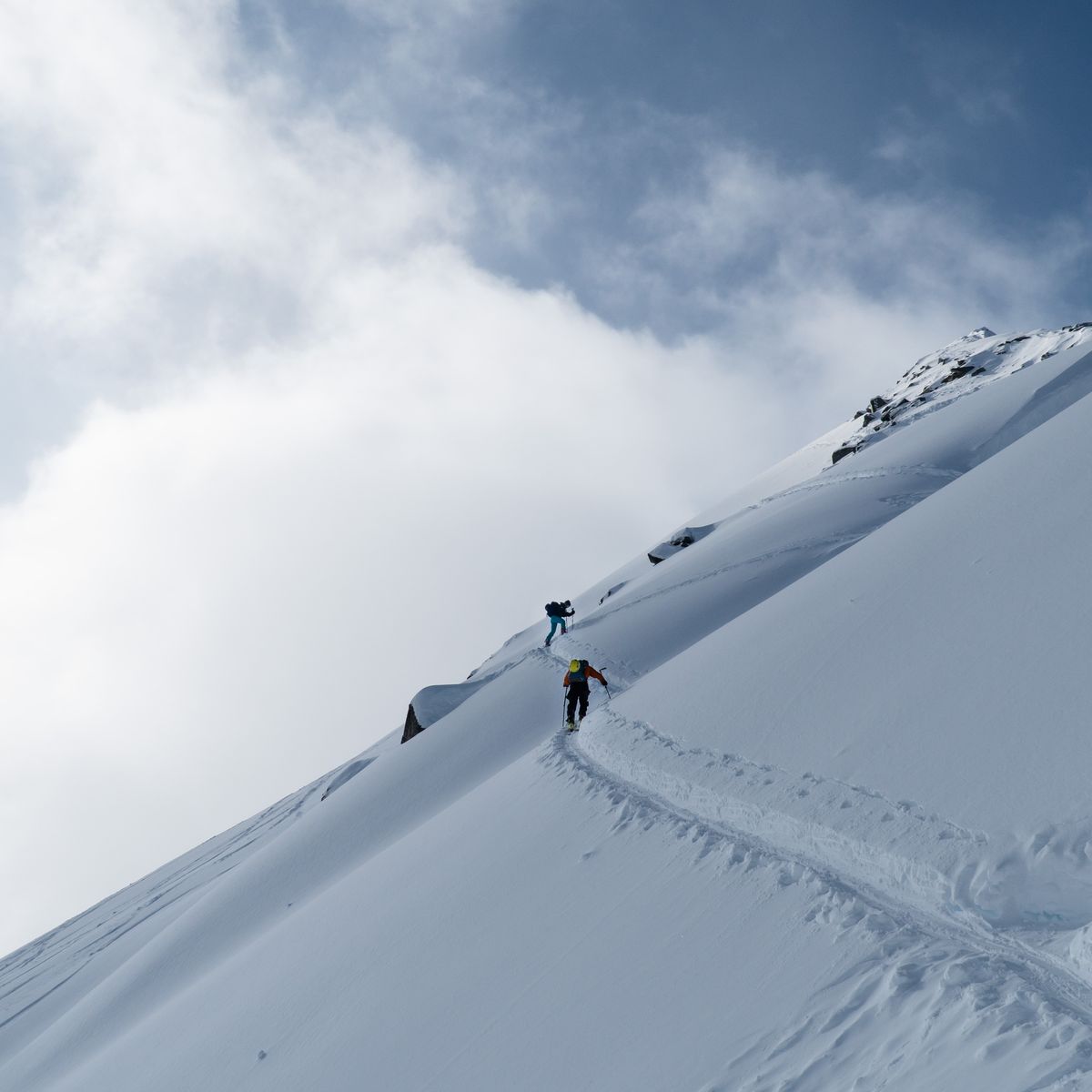 Two climbers ascend a steep, snow-covered mountain slope under a partly cloudy sky, leaving a trail behind them.