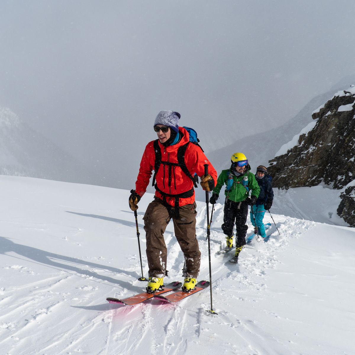 Three skiers in colorful gear ascend a snowy mountain trail, with rocky cliffs and a misty sky in the background.