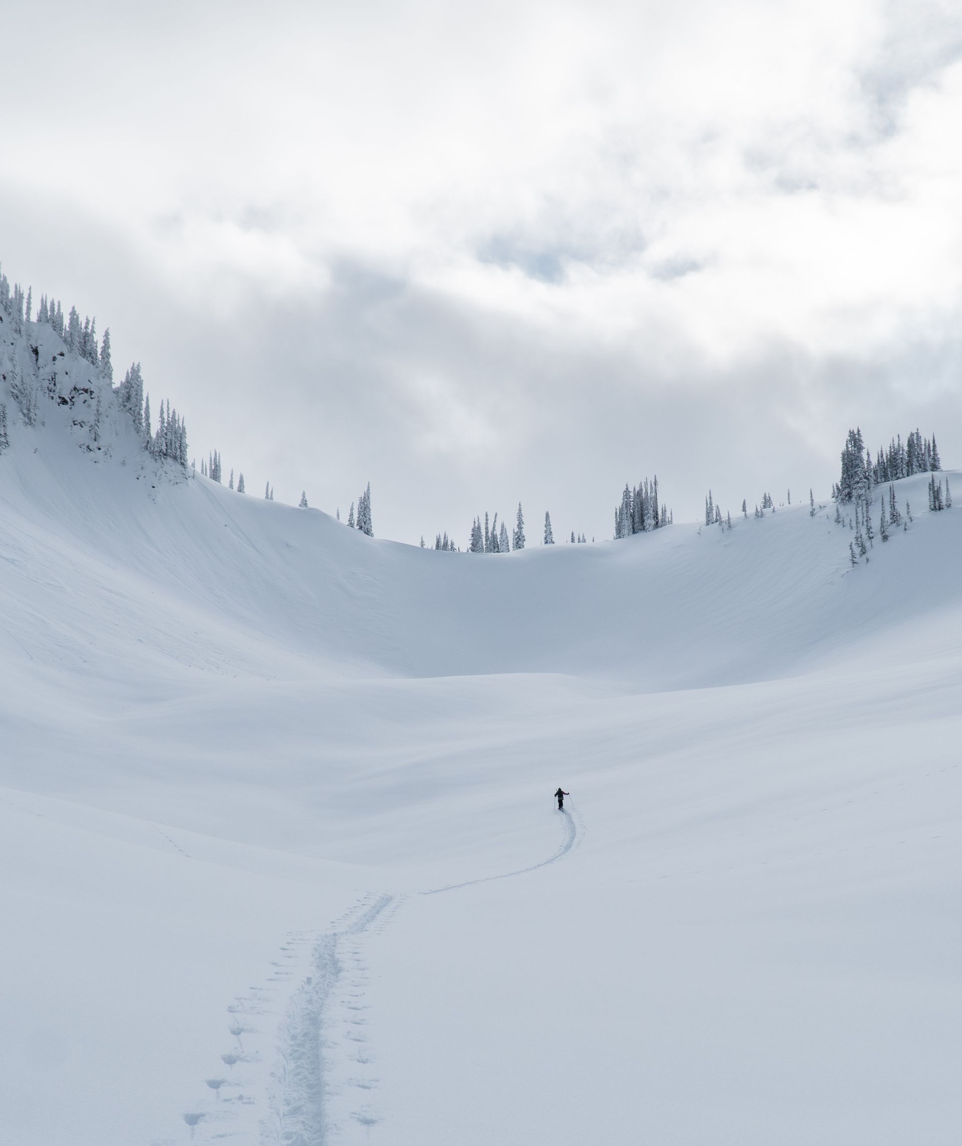 A lone skier traverses a vast, snow-covered mountain landscape under a cloudy sky, leaving a single trail behind.