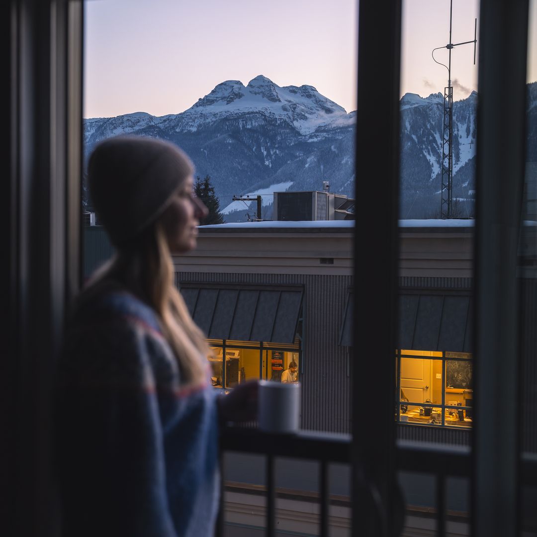 A person in a beanie looks out a window at snowy mountains during sunset, holding a mug. A lit room is visible in a building opposite.