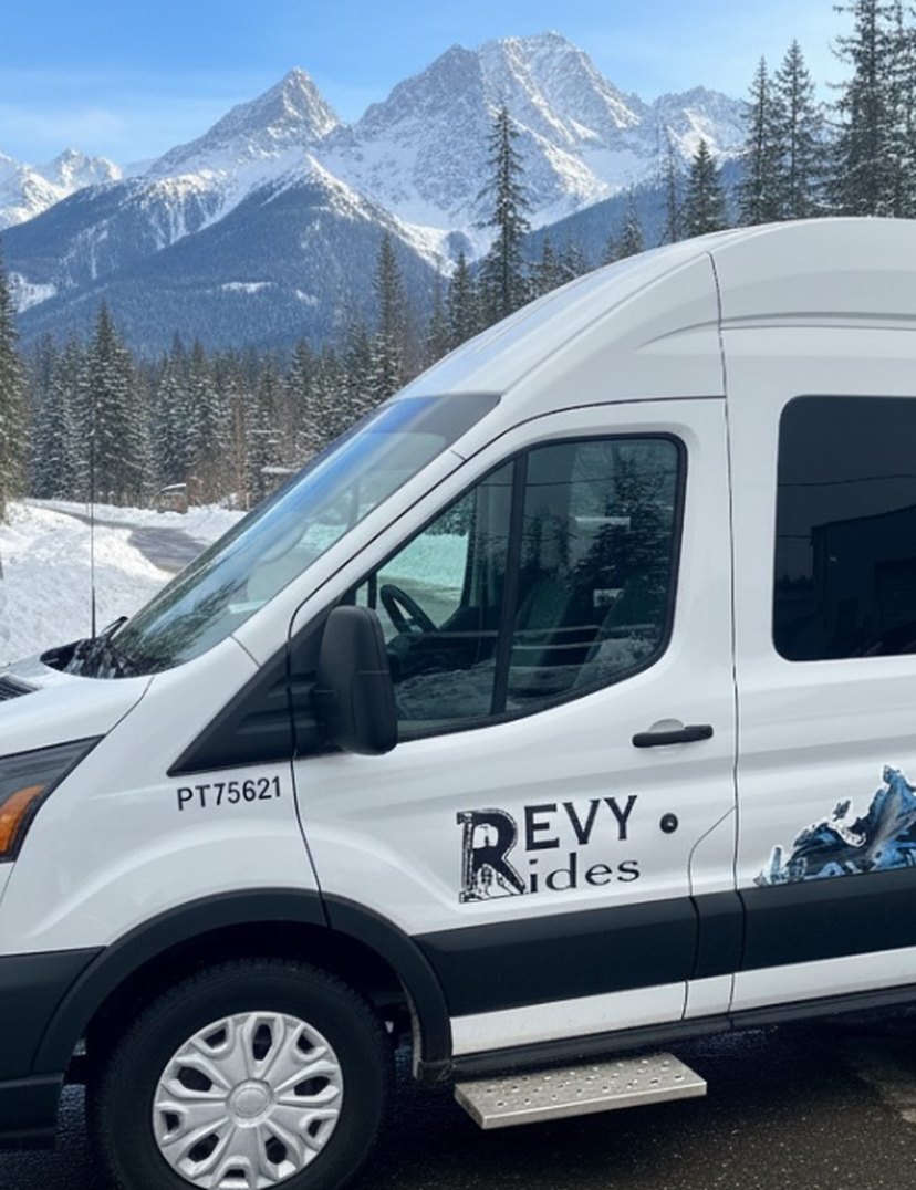 White "Revy Rides" passenger van parked on a snowy mountain road with pine trees and towering snow‑capped peaks behind.