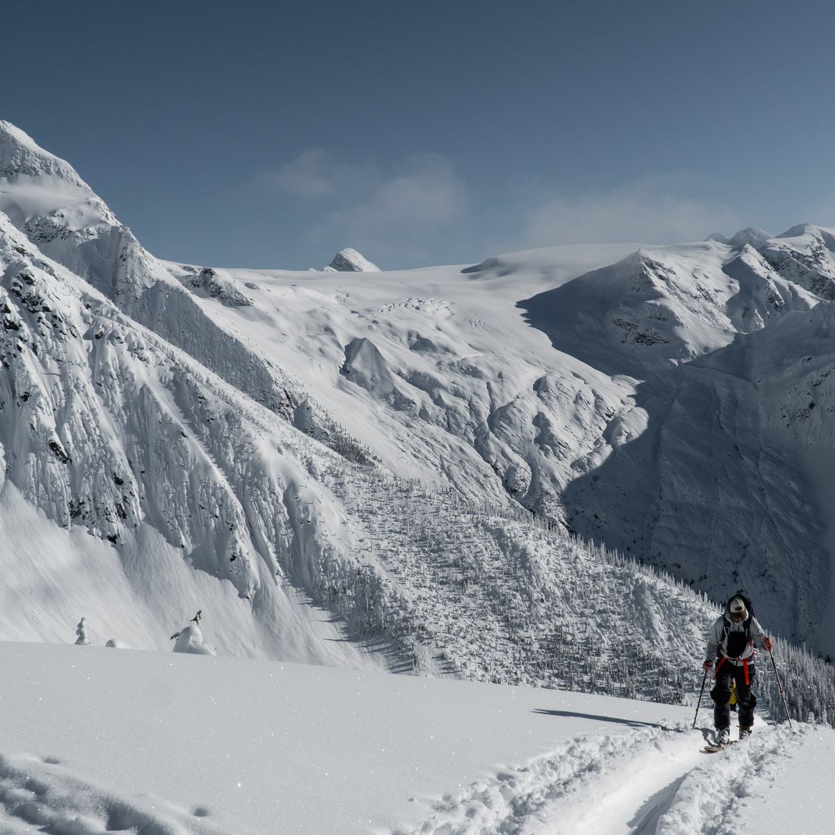 A person hikes up a snowy mountain trail surrounded by vast, rugged snow-covered peaks under a clear blue sky.