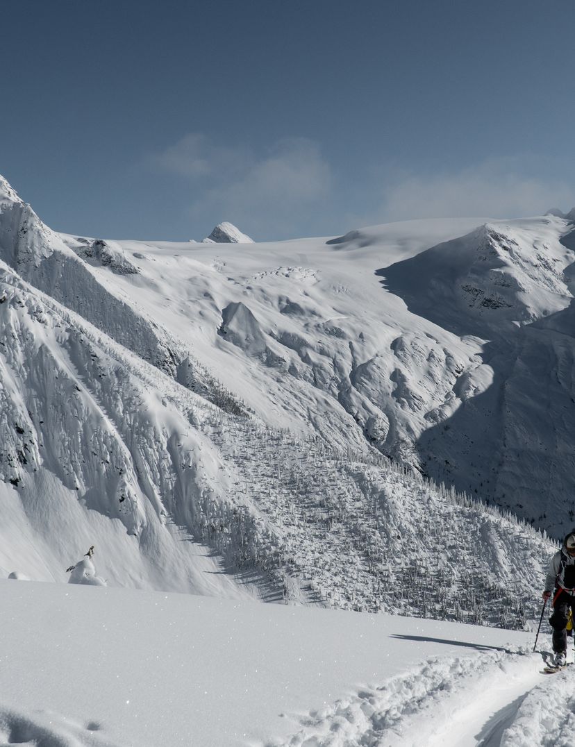 A person with a backpack hikes up a snowy mountain trail, surrounded by vast snow-covered peaks under a clear blue sky.