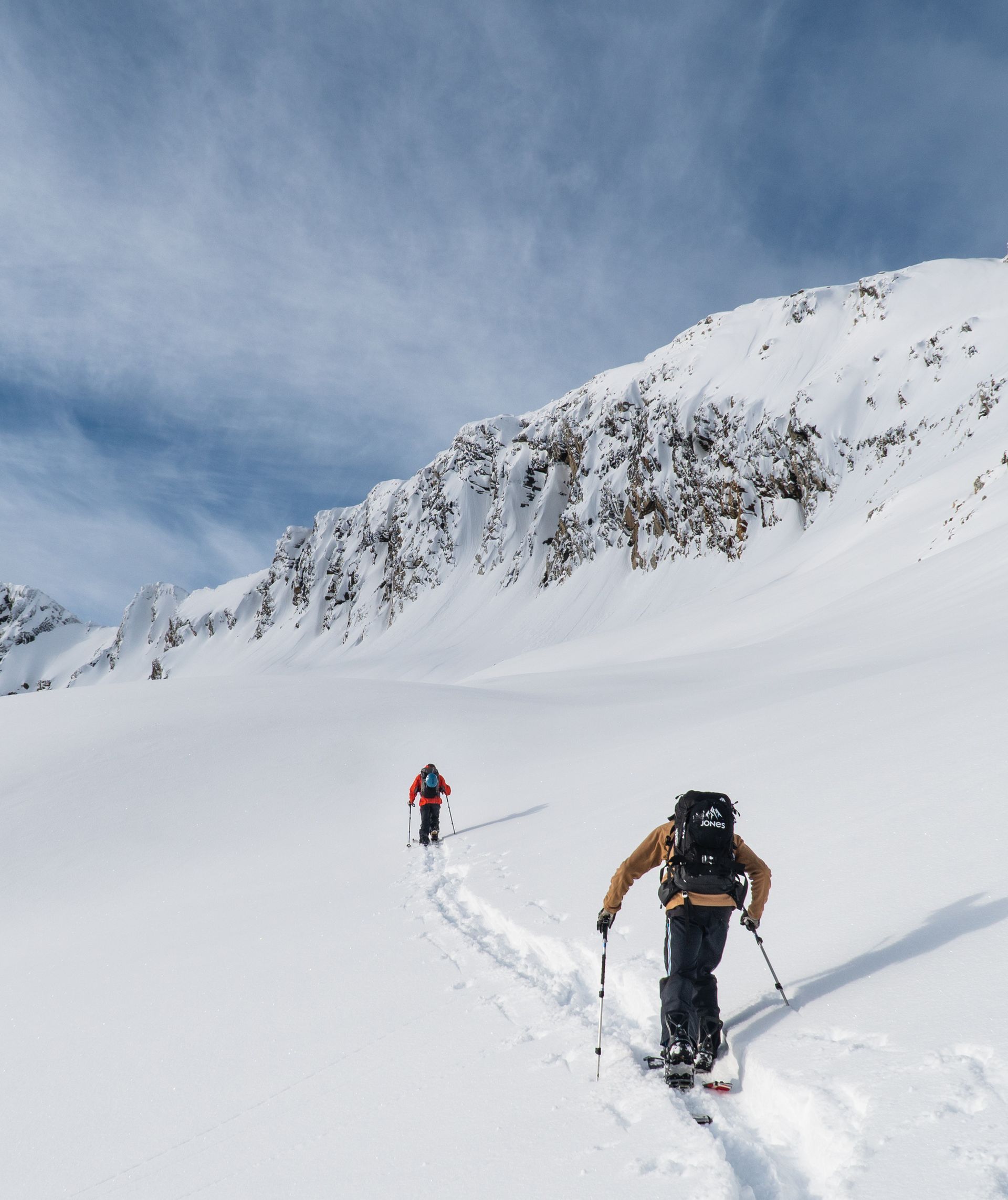 Two skiers ascend a snowy mountain slope under a partly cloudy sky, leaving tracks behind them. Rocky peaks are visible in the background.