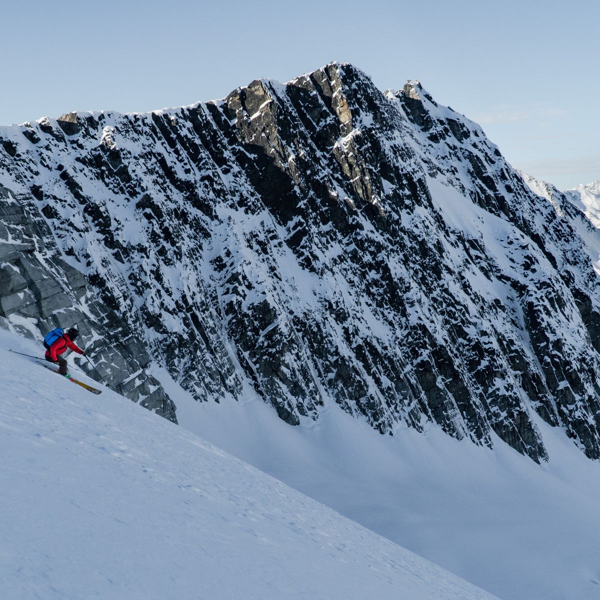 A skier in a red jacket descends a steep, snow-covered mountain slope with rugged, rocky peaks in the background under a clear sky.