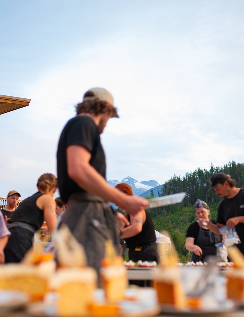 People preparing food outdoors with a mountain view in the background, under a striped awning near a wooden building.