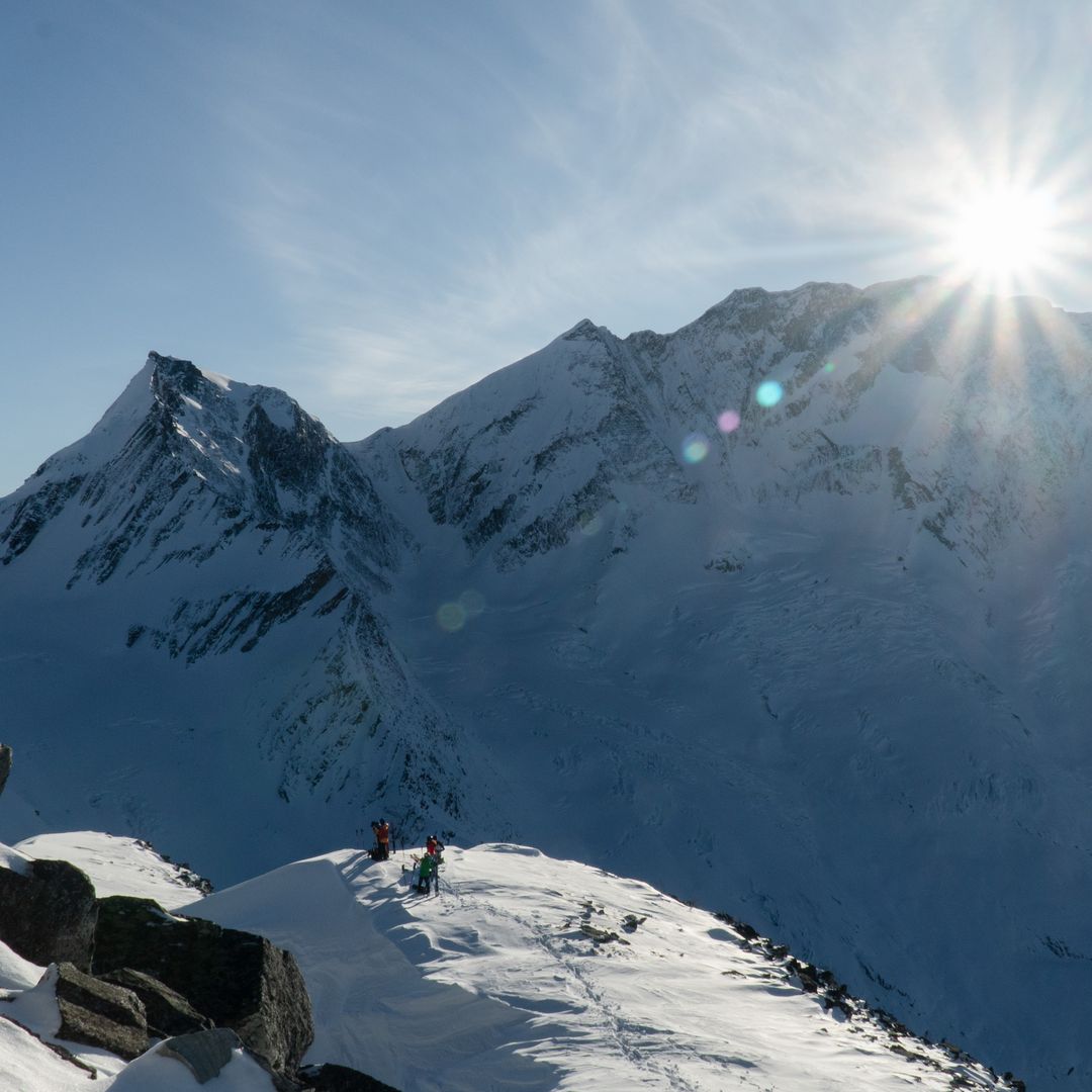 Snowy mountain landscape with two climbers on a ridge, sun shining brightly over distant peaks under a clear blue sky.