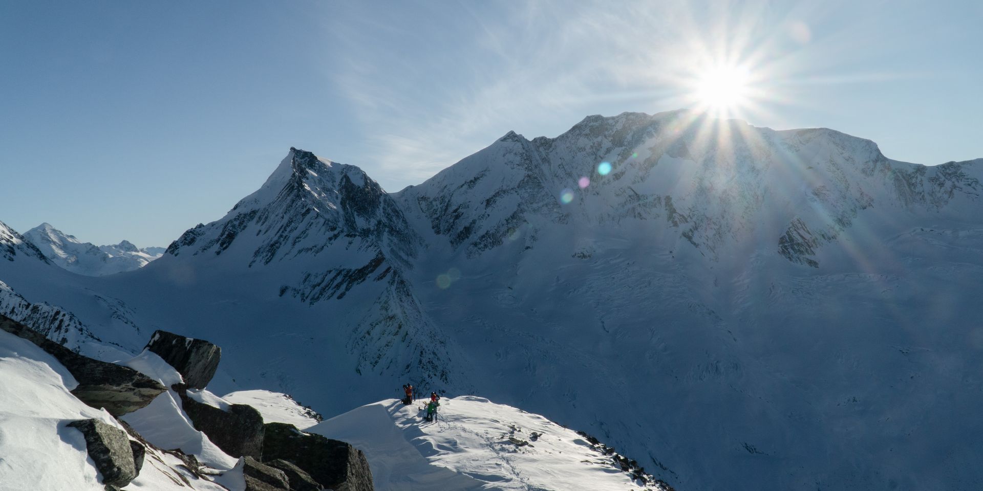 Snowy mountain landscape with two climbers on a ridge, sun shining brightly over distant peaks under a clear blue sky.