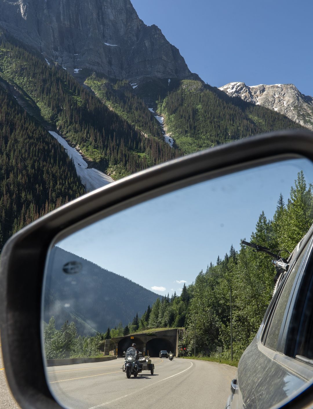Car side mirror reflecting a mountain road scene with a motorcyclist approaching a tunnel, surrounded by lush green trees and rocky peaks.