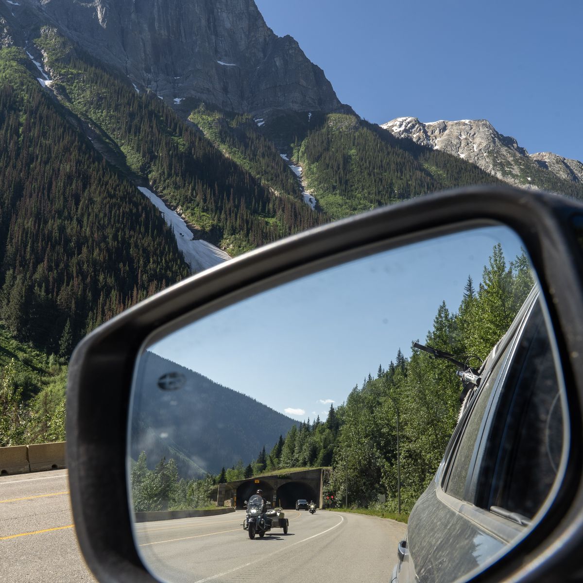 Car side mirror reflecting a mountain road scene with a motorcyclist approaching a tunnel, surrounded by lush green trees and rocky peaks.