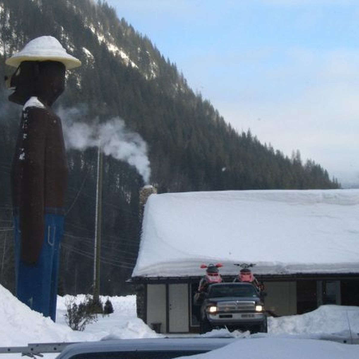 Large statue of a person in a hat by a snow-covered building and truck, set against a mountainous, snowy landscape.