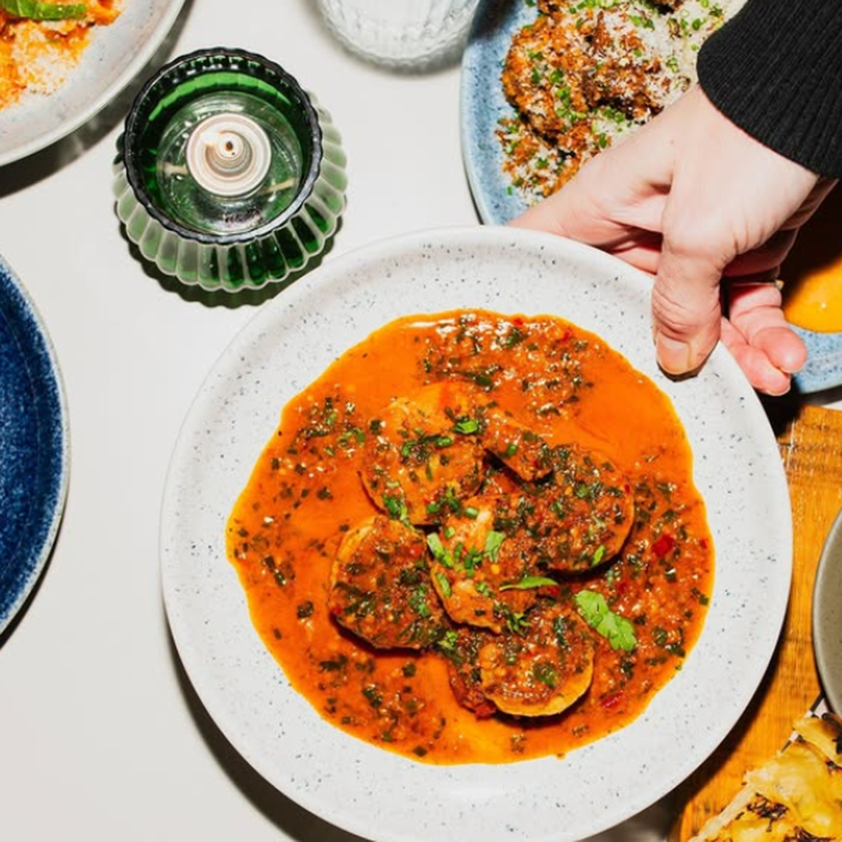 A hand placing a white plate of ravioli in rich tomato sauce on a table, surrounded by other dishes and a green candle holder.