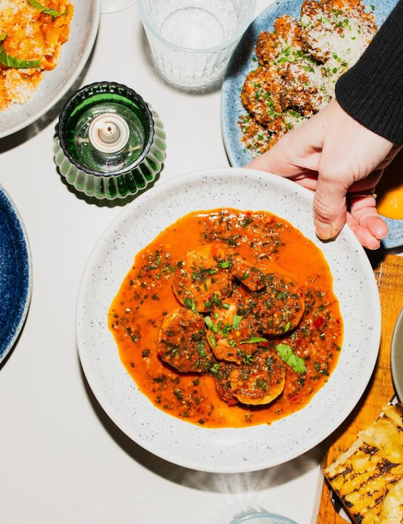 A hand placing a white plate of ravioli in rich tomato sauce on a table, surrounded by other dishes and a green candle holder.
