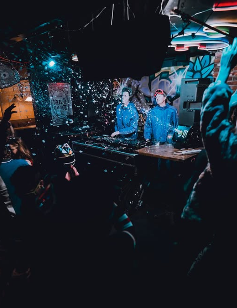 Two DJs in blue hoodies play amid confetti and neon lights as a crowd raises hands in a graffiti-filled club