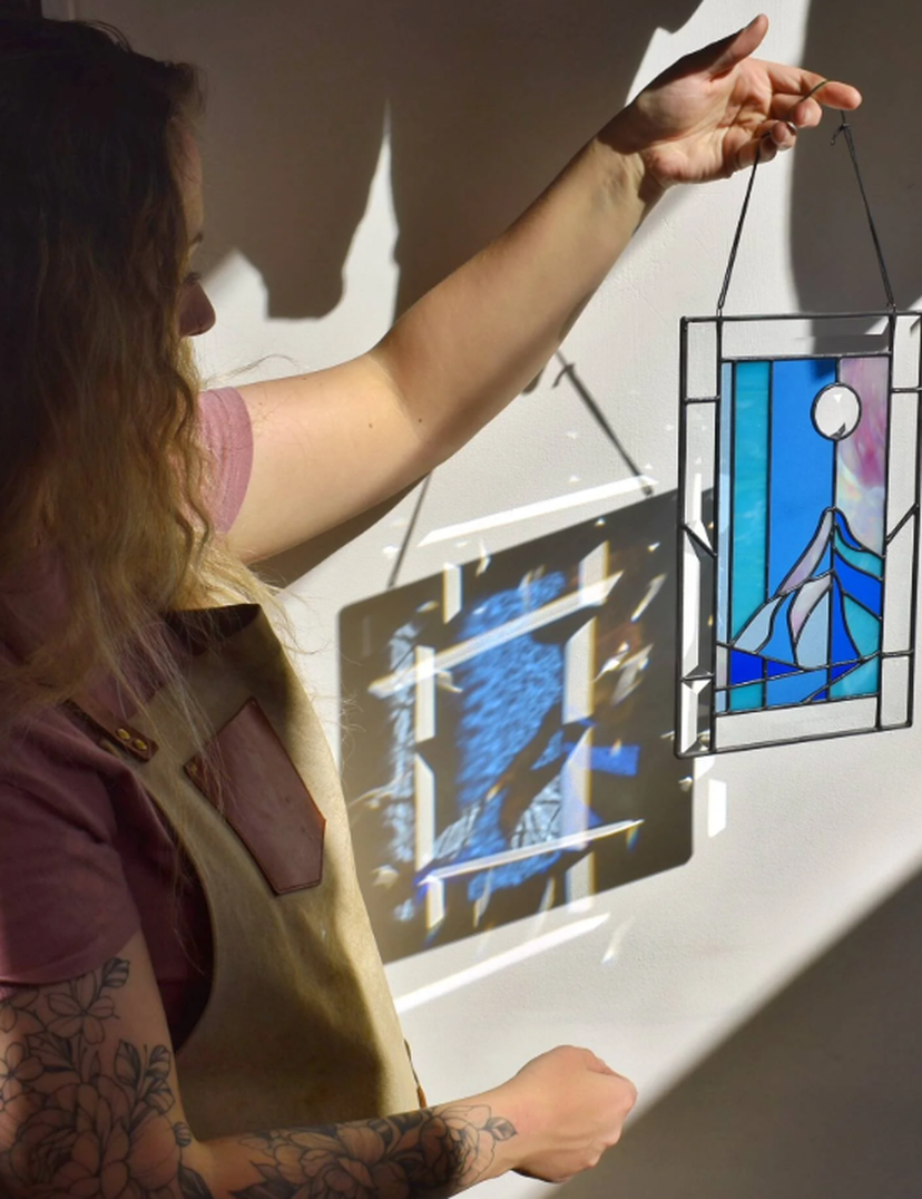 Person holding a stained glass panel depicting a mountain and moon, casting a colorful shadow on the wall.