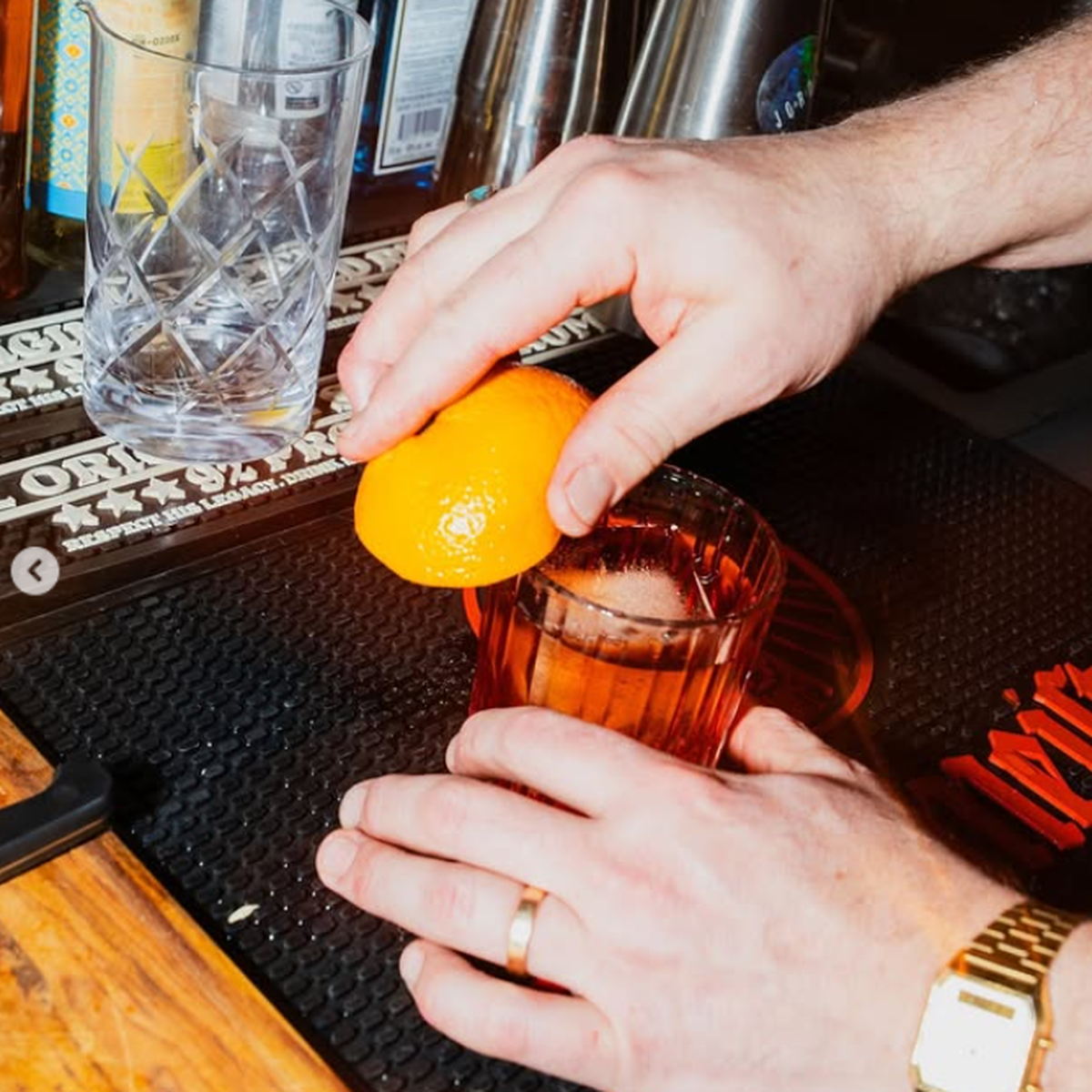 Person garnishing a cocktail with an orange peel at a bar, with various bar tools and bottles in the background.