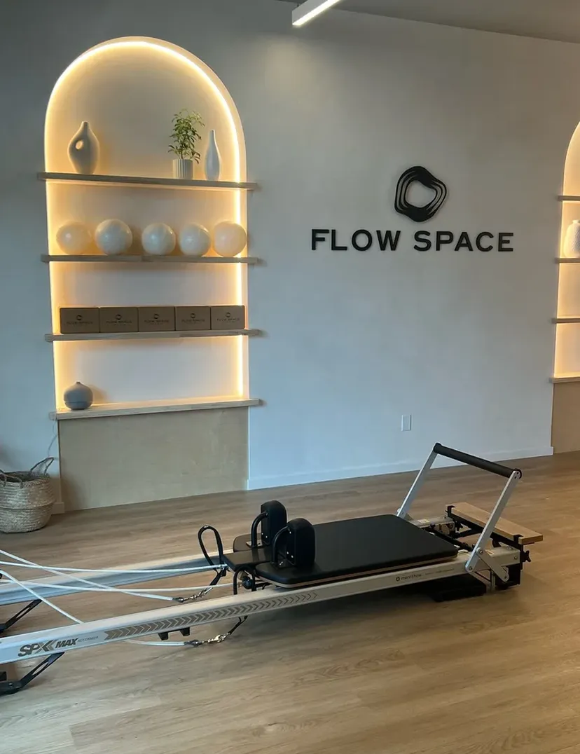 Pilates studio with a reformer machine, wall shelves with vases and boxes, and "FLOW SPACE" logo on the wall.