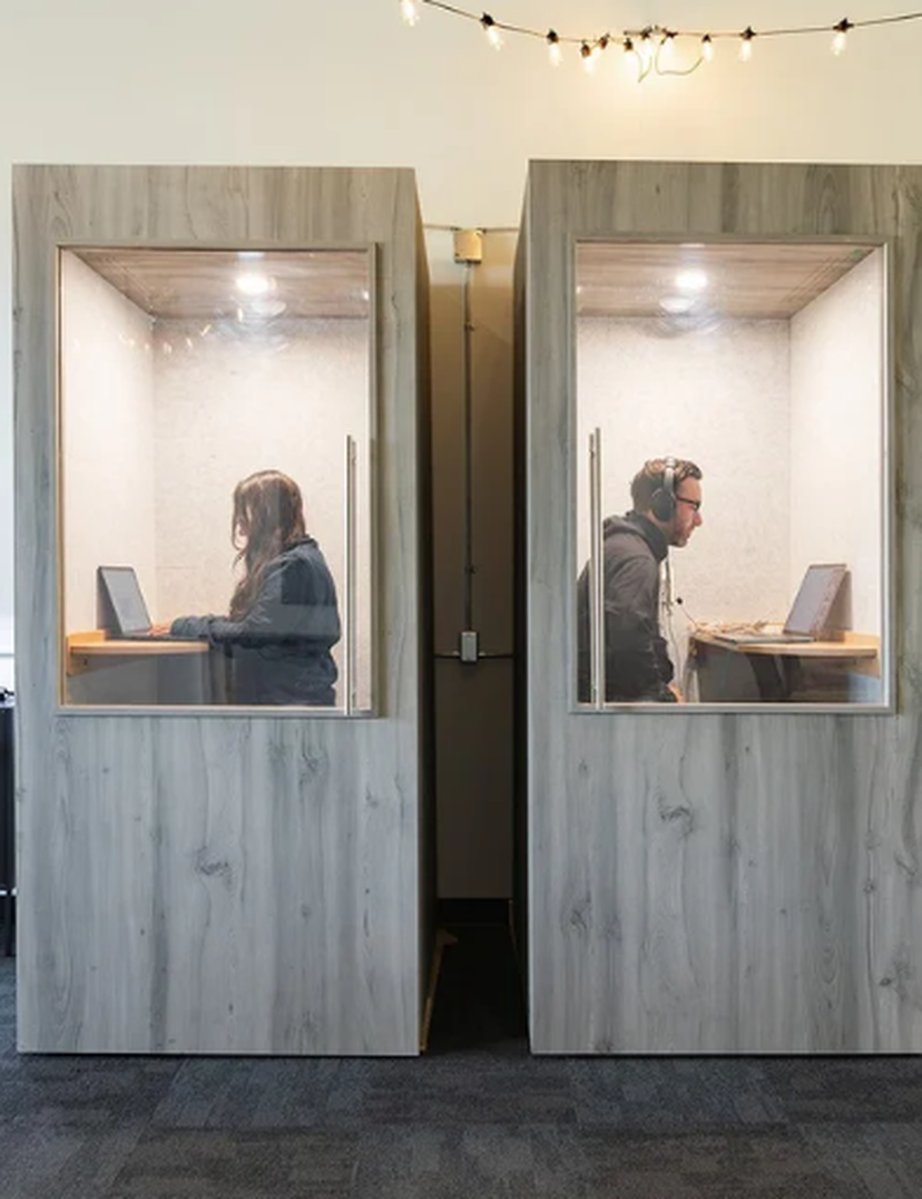Two people working in adjacent, soundproof booths with laptops, separated by a wall. Office chairs and shelves are visible in the background.