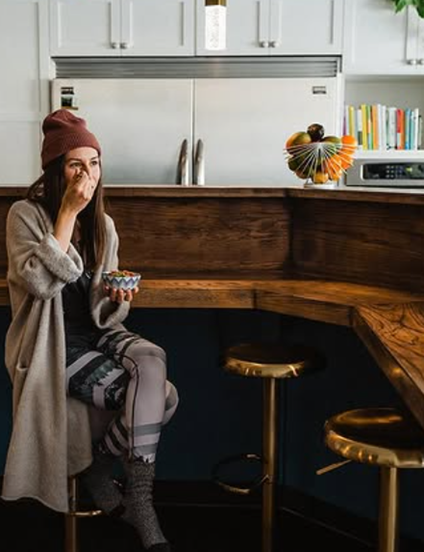 Woman in cozy attire eats from a bowl, sitting on a stool in a modern kitchen with wooden counters and brass accents.
